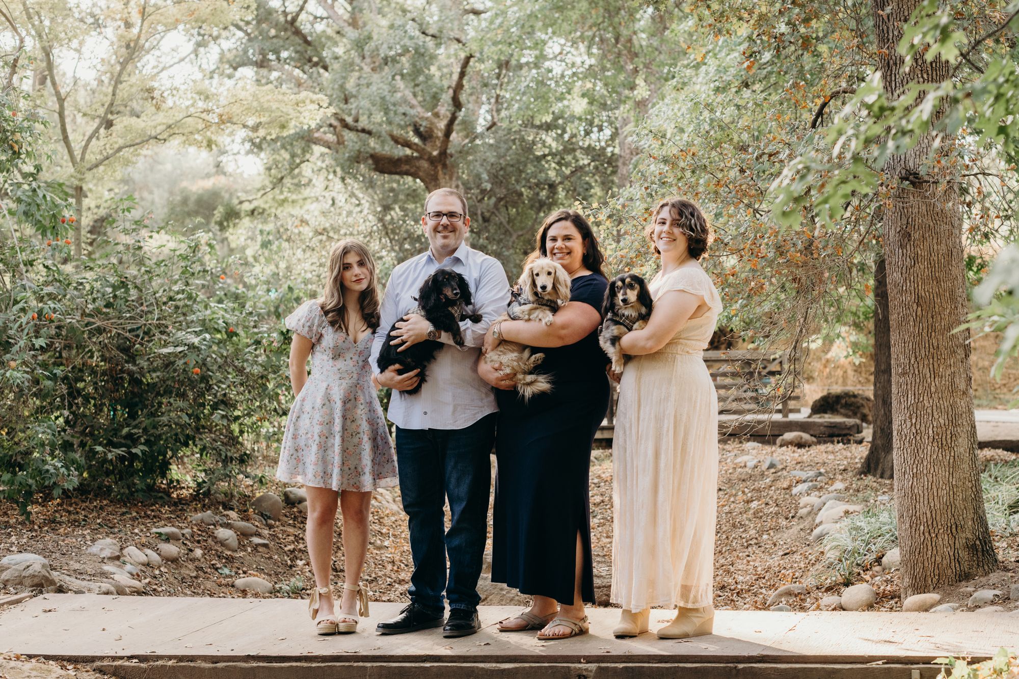 Jessica with her husband Kevin, their two daughters, and three long-haired dachshunds on a wooden footbridge under the oaks